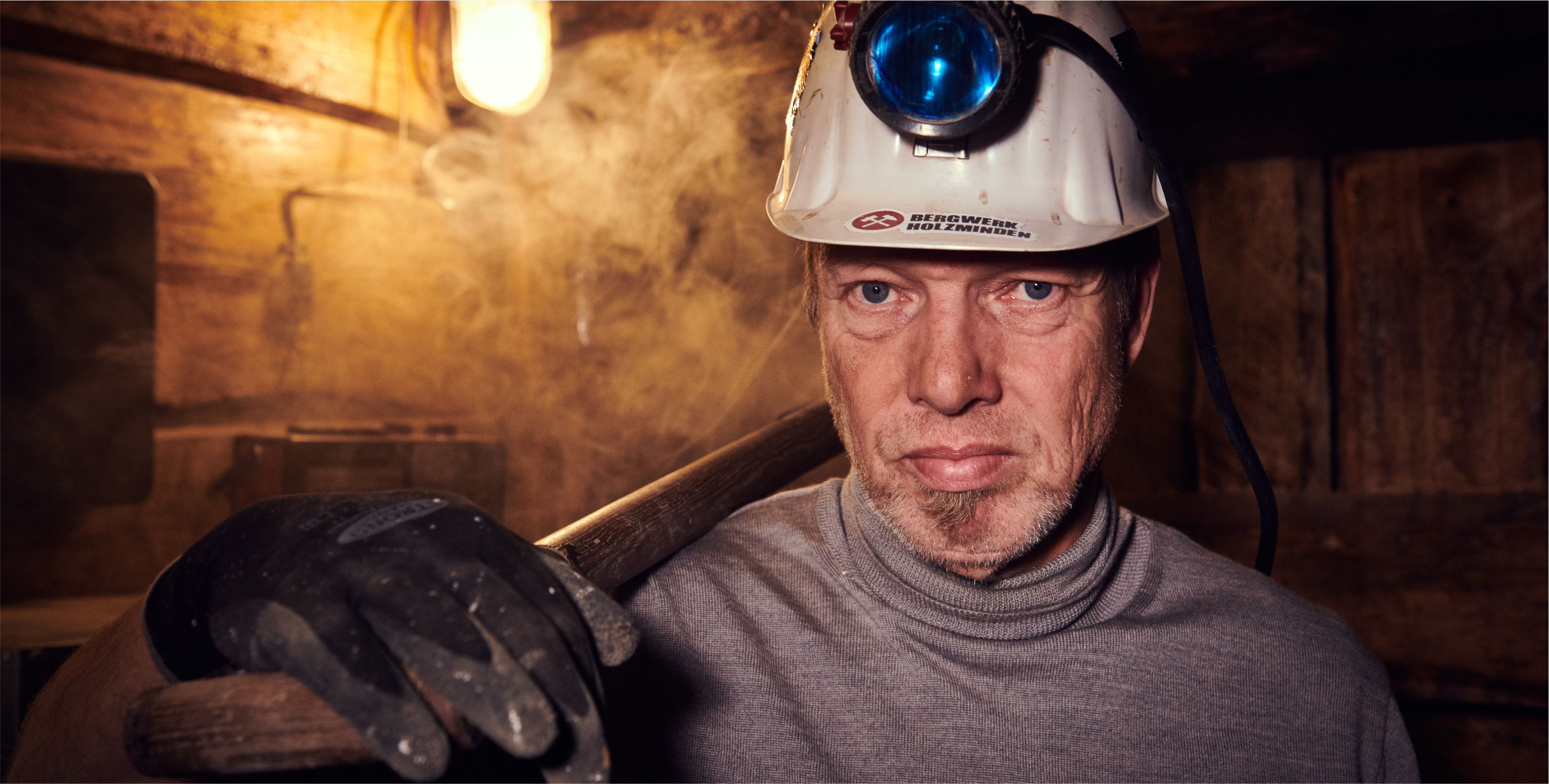 Ein Mann mit Helm und Spaten ber der Schulter im Bergwerk blickt im Halbportrt in die Kamera